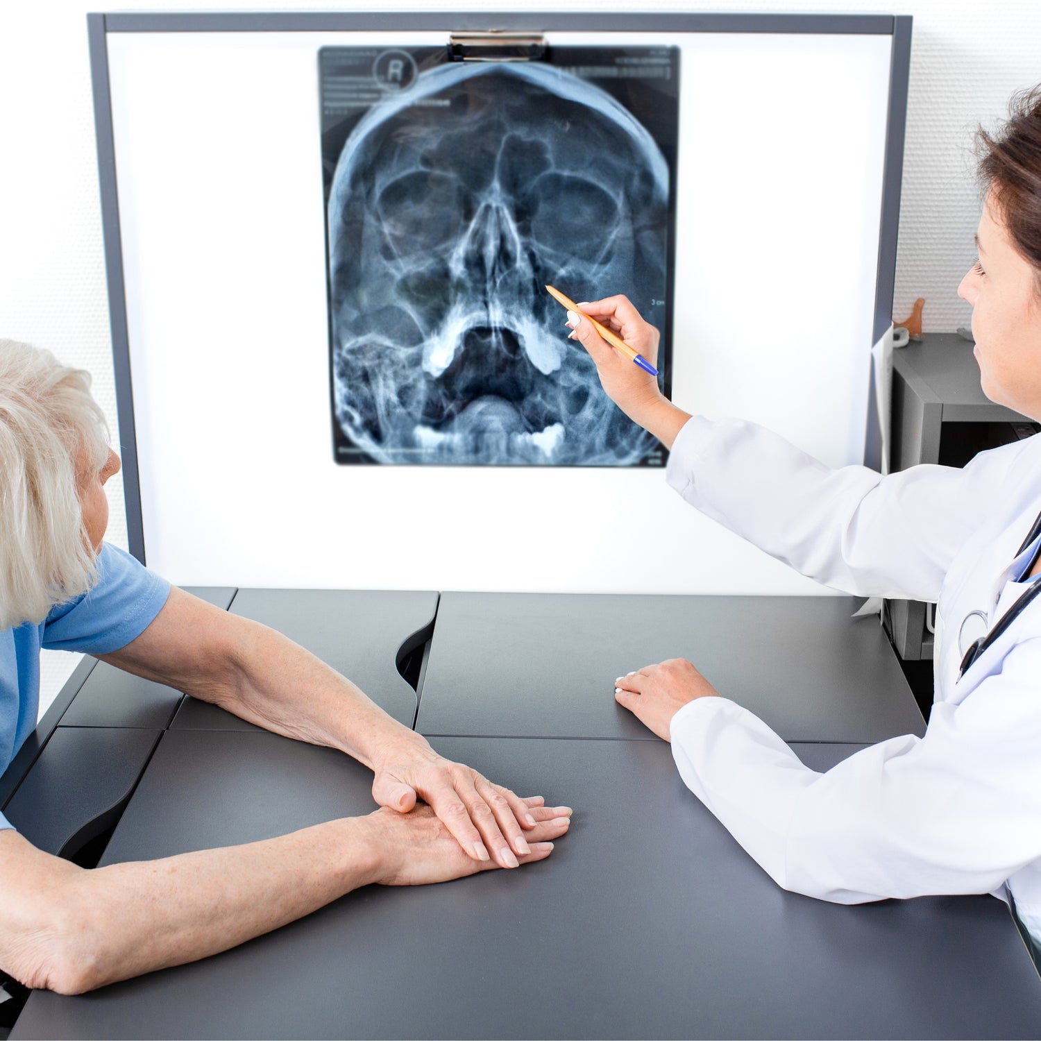 Alte Frau sitzt gegenüber von einer Ärztin. Die Ärztin deutet mit einem Stift auf die Röntgenaufnahme eines Schädels. Beide schauen auf das Bild.